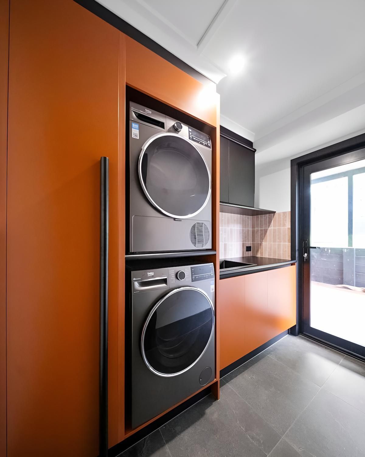 Orange cabinetry encloses a stacked washer and dryer. A dark countertop and a glass door are visible — CK Cabinetry in Coolum Beach, QLD