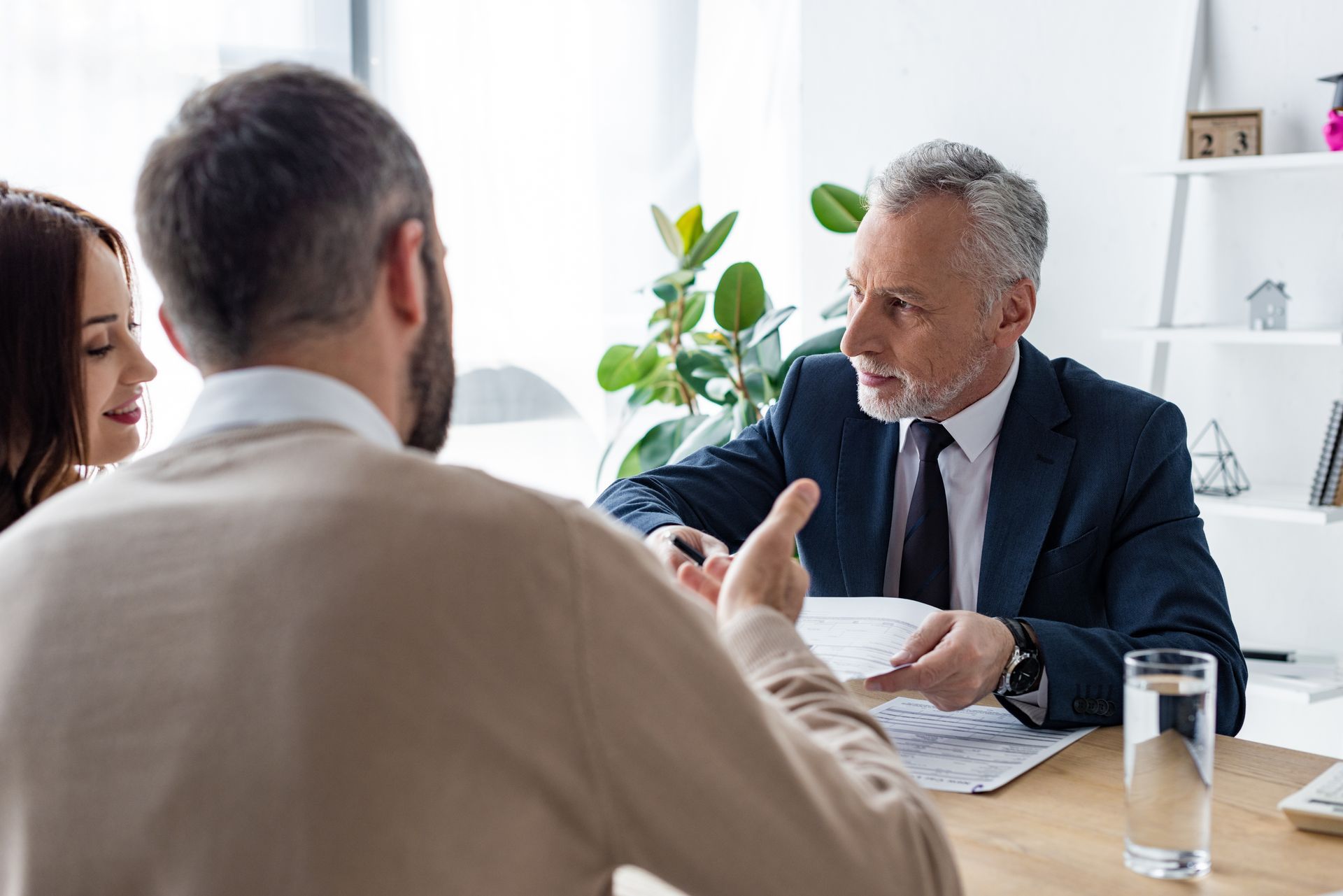 A man in a suit gestures at paperwork while consulting a couple at a desk.