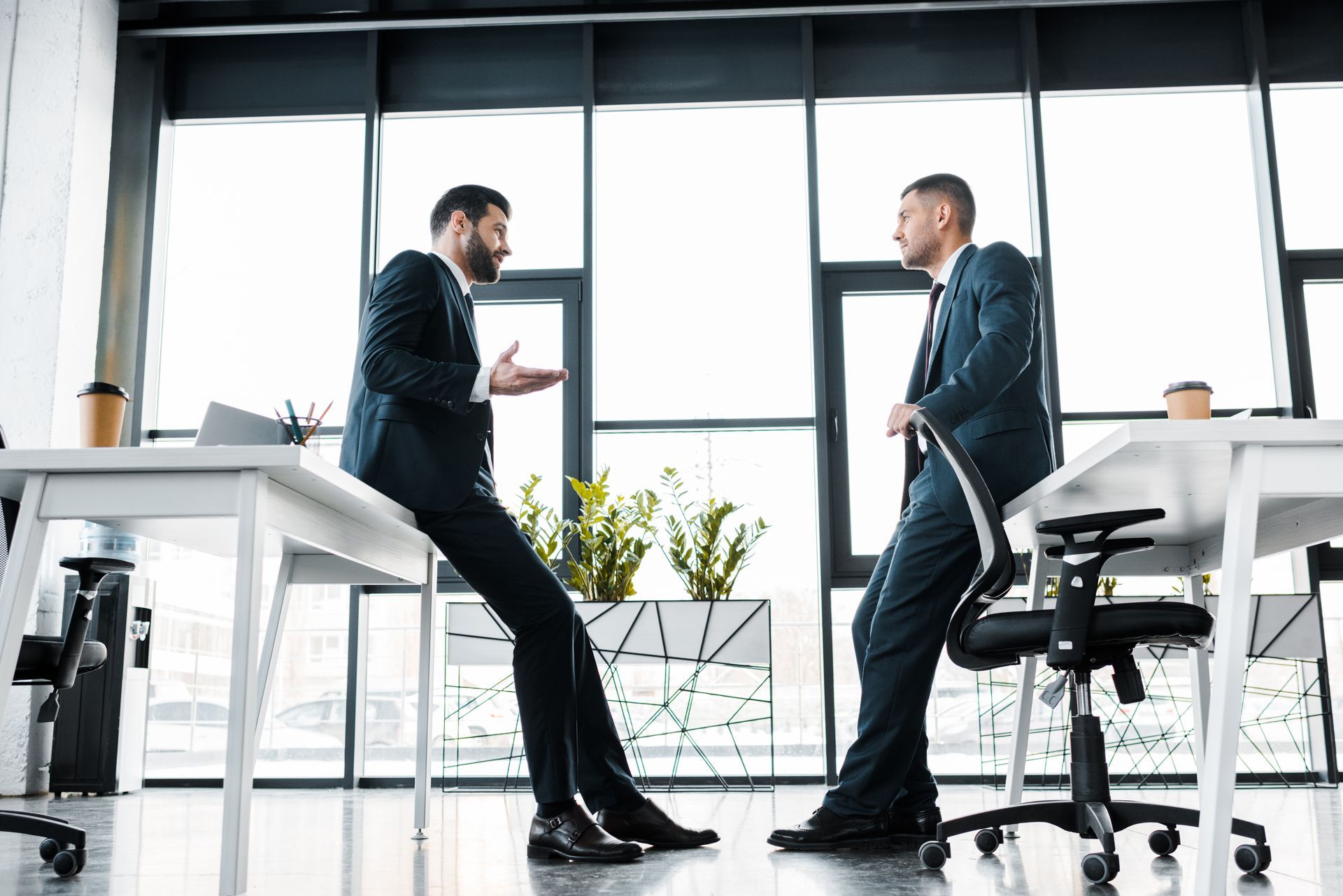 Two men in suits converse in an office, one leaning on a desk, the other on a chair, near a window.