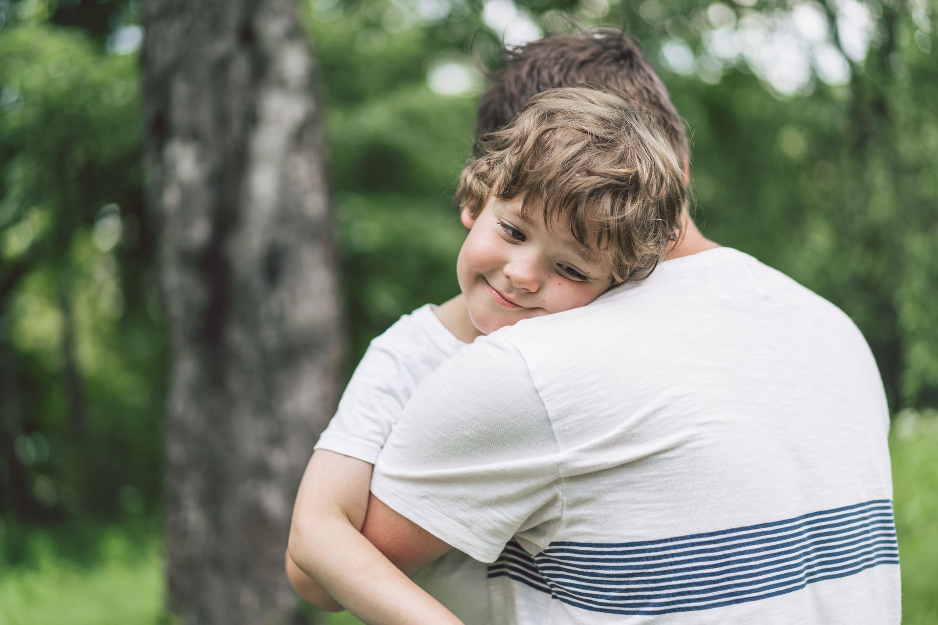 Boy hugging a person, resting head on shoulder. Outdoors, near a tree.