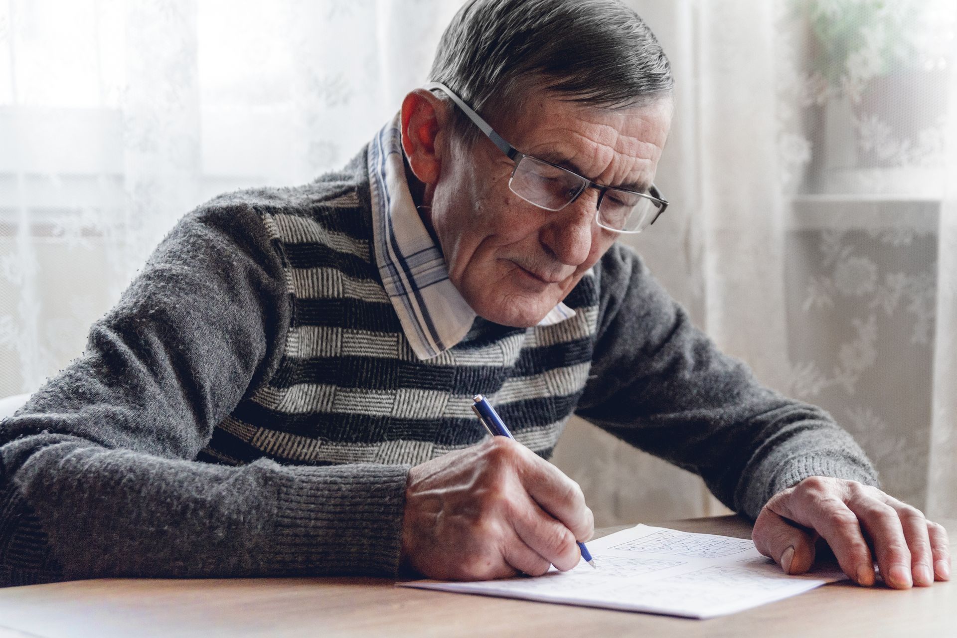 Older person with glasses writes at a table indoors.