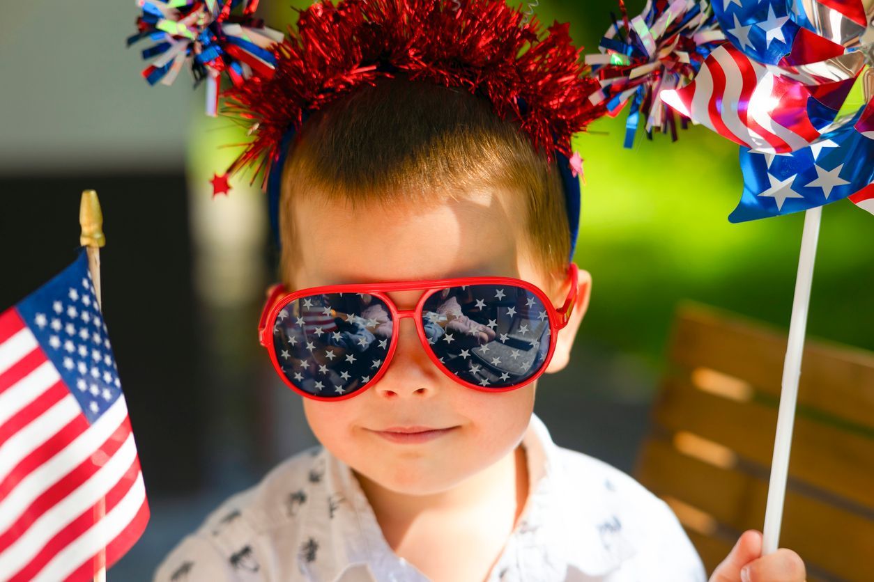 Child in red sunglasses and patriotic headband, holding small U.S. flags.