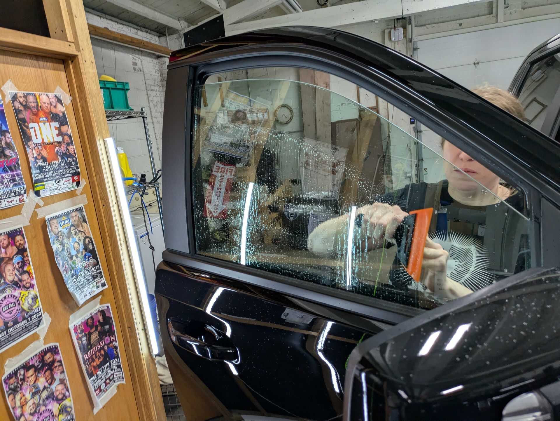A man is applying window tinting to a black car in a garage.