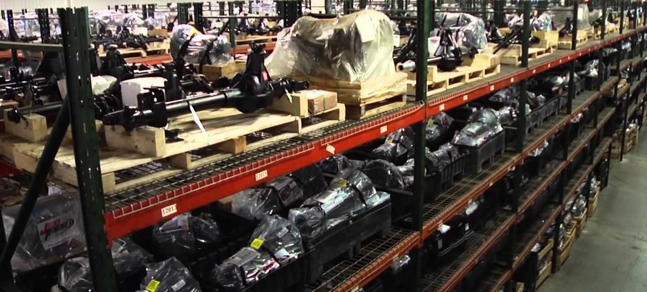 Warehouse shelves filled with car parts, some on wooden pallets. The parts are wrapped in plastic.