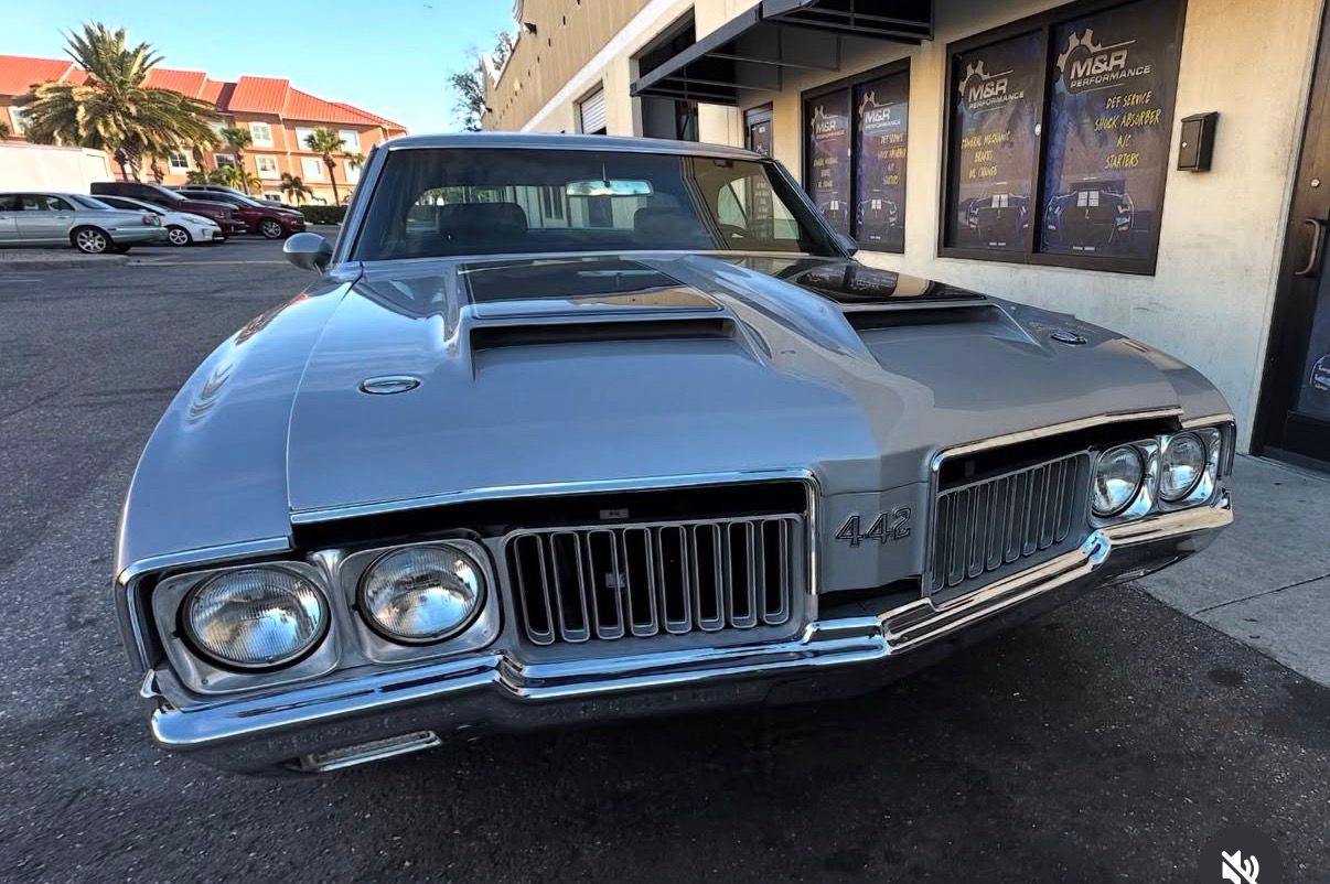 Silver 1970 Oldsmobile 442, front view, parked near a building with awnings, sunny outdoor setting.