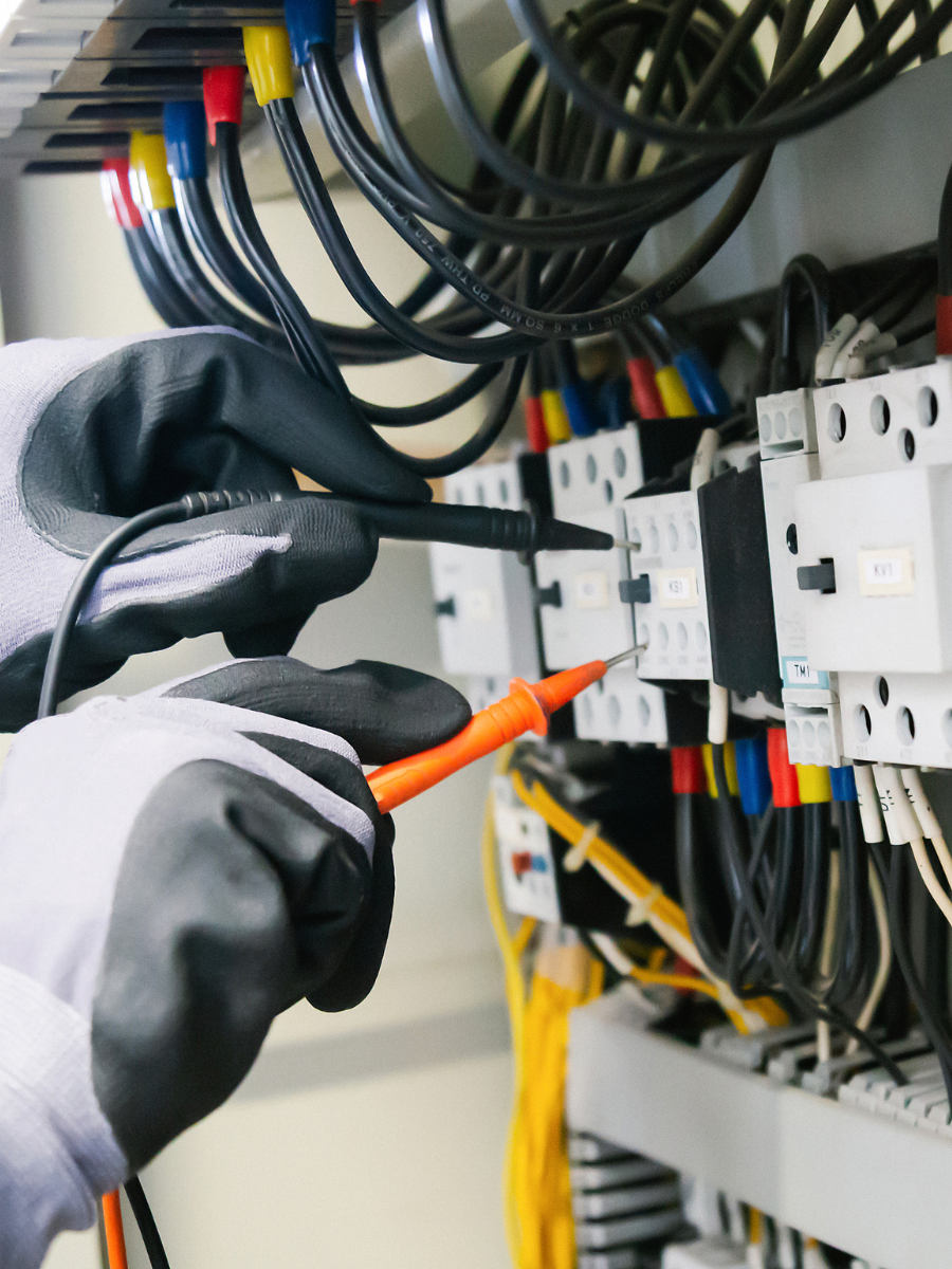 Electrician testing electrical panel with a multimeter; gloved hands, colorful wires.