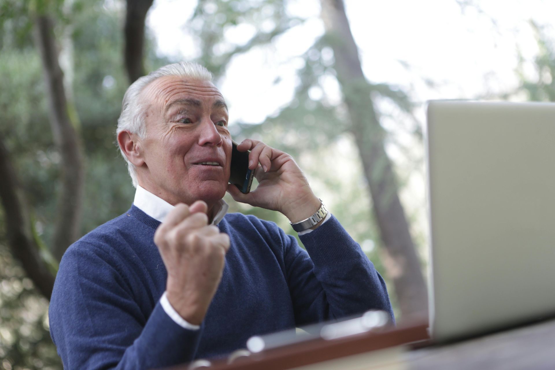 An elderly man is talking on a cell phone in front of a laptop computer.