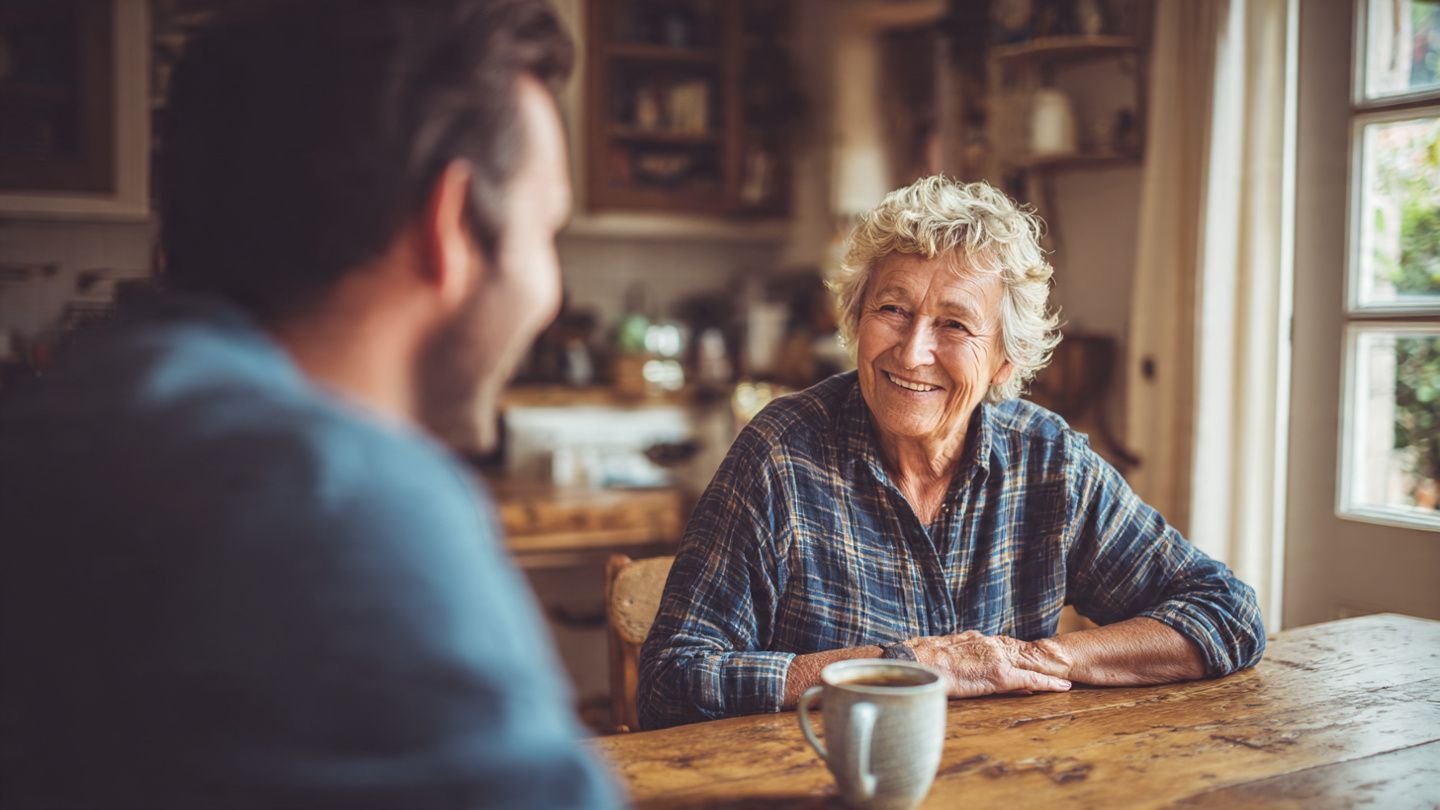 Two people talking at a rustic wooden table with a coffee mug in a cozy kitchen.