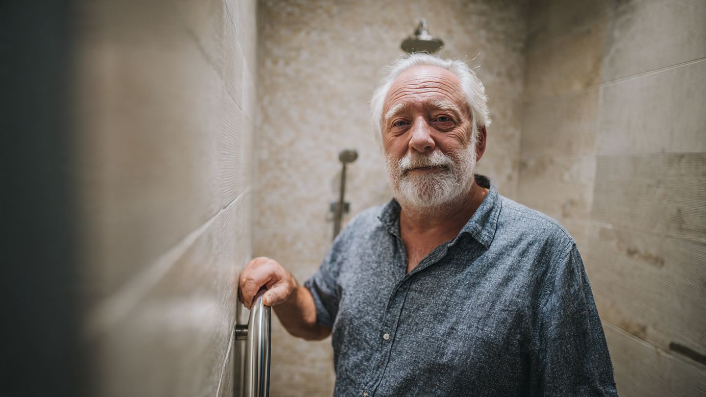 A man with a grey beard wearing a blue patterned shirt stands in a tiled shower, resting his hand on a grab bar.