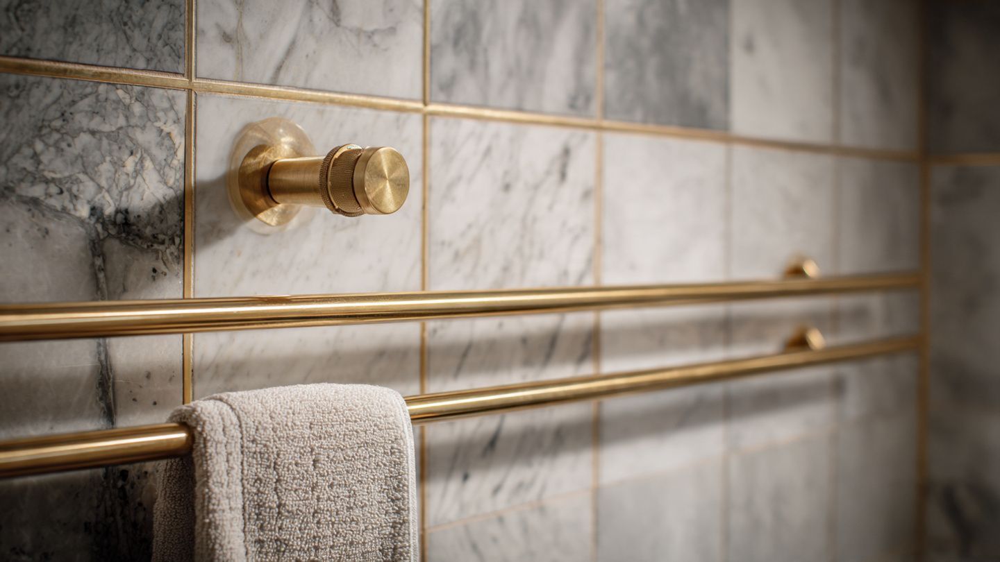 A gold towel bar and a separate matching hook are mounted on a gray and white marble tiled wall with a hanging towel.