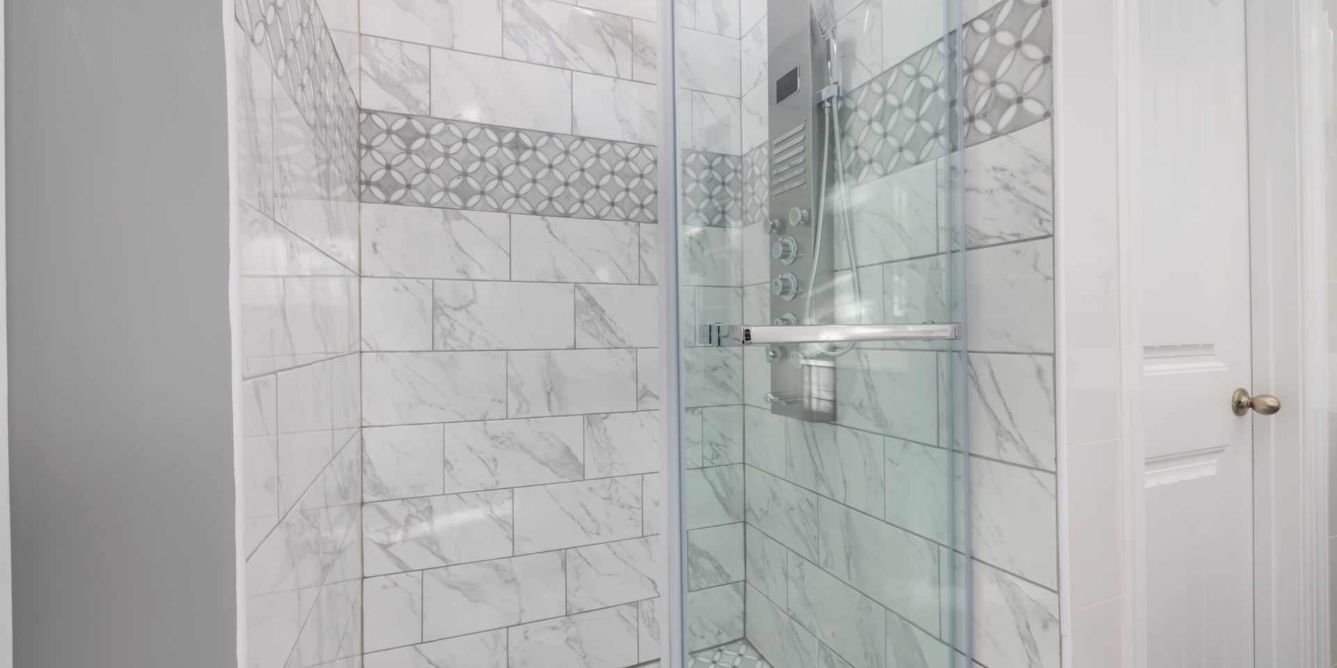 A marble-tiled shower with a glass door, a white door, and a silver shower head.