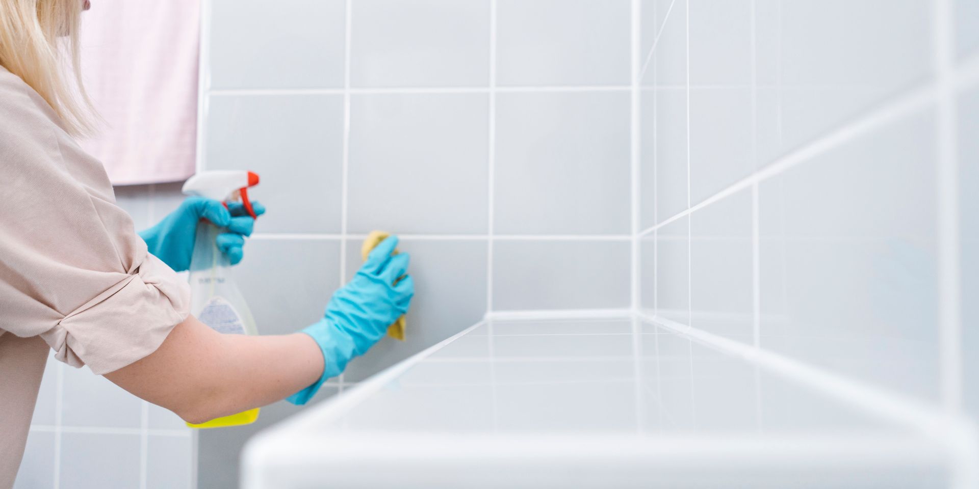 Person cleaning a bathroom with spray bottle and sponge. Wearing blue gloves, light-colored tiles.