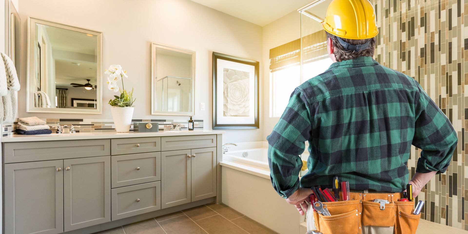 A construction worker in a bathroom, wearing a hard hat and tool belt, surveys the space.