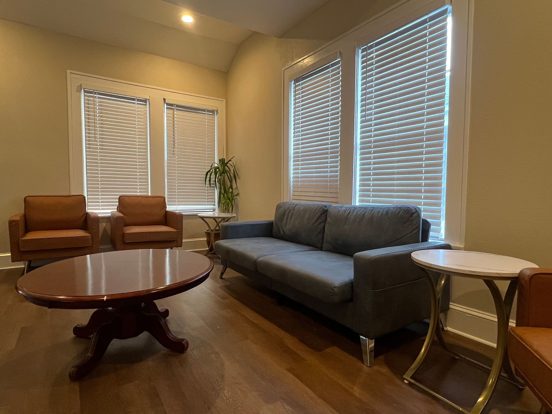 Living room with brown hardwood floors, gray sofa, two chairs, round coffee table, and windows with blinds.