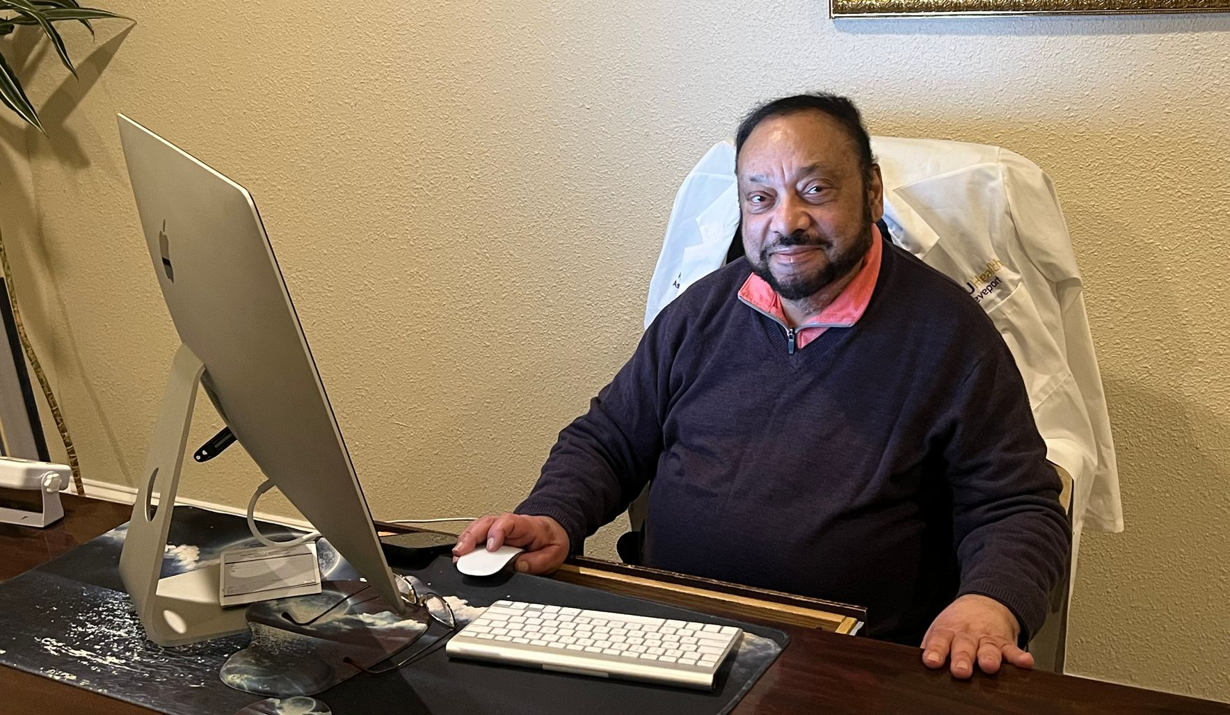 Dr. Ibrahim sitting at desk with computer, looking at camera. Brown sweater, beige wall, white lab coat in background.