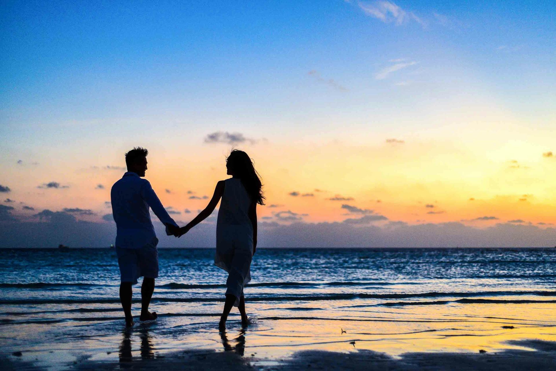 Couple holding hands, silhouetted on a beach, watching a sunset over the ocean.