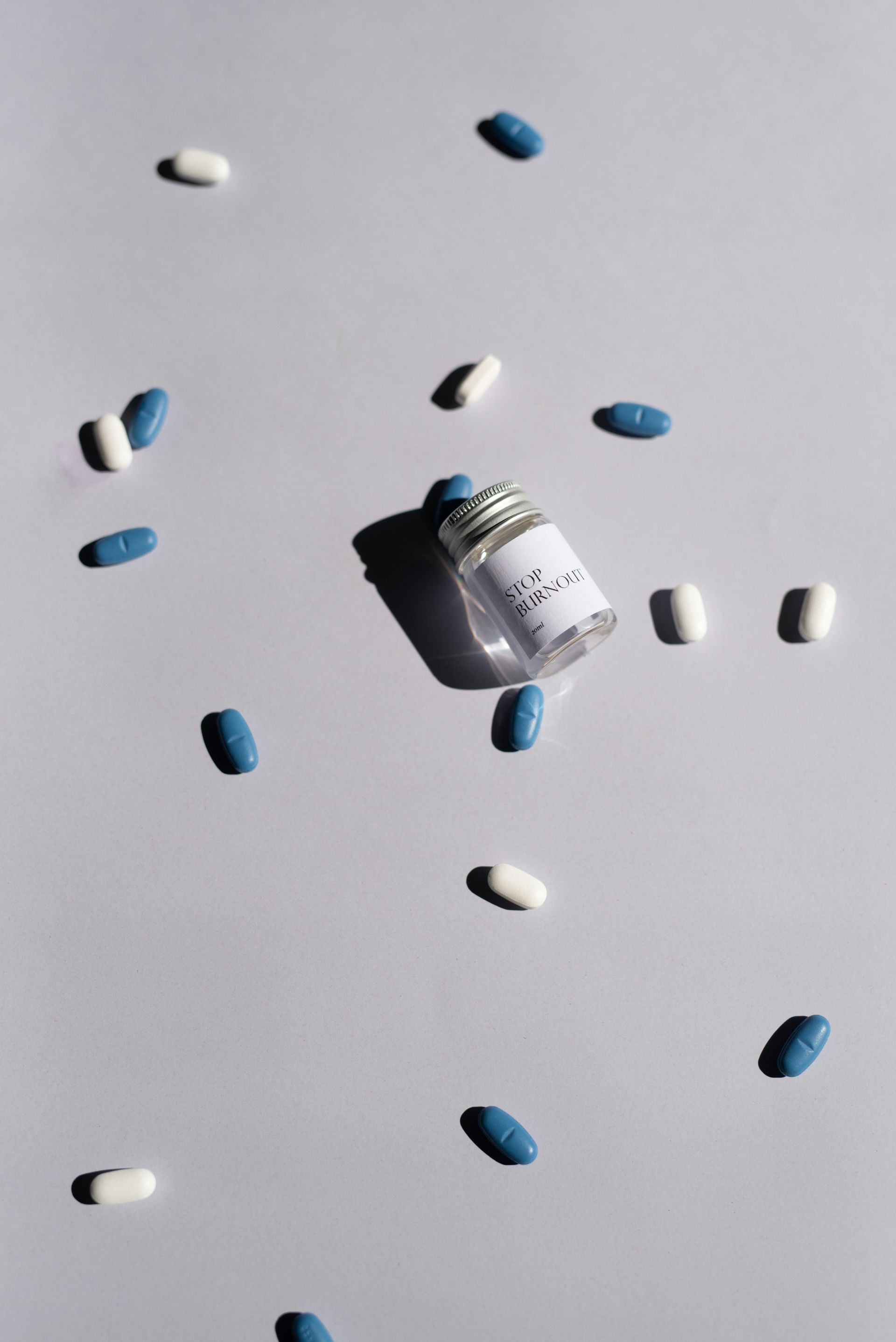 Scattered blue and white pills surround a small glass bottle with a label, casting shadows on a white surface.