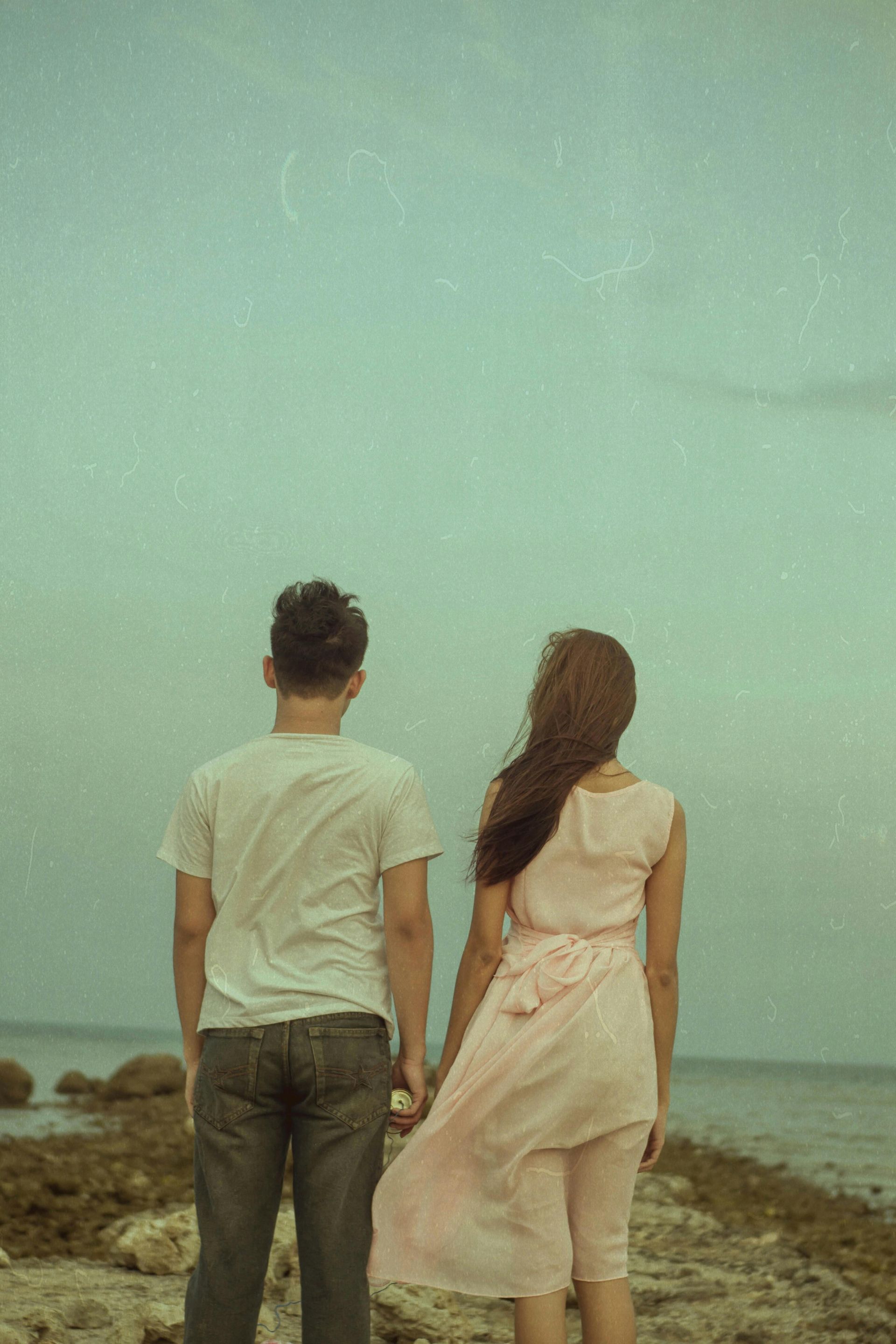 Couple standing on a rocky shore, looking out at the ocean under a cloudy sky.