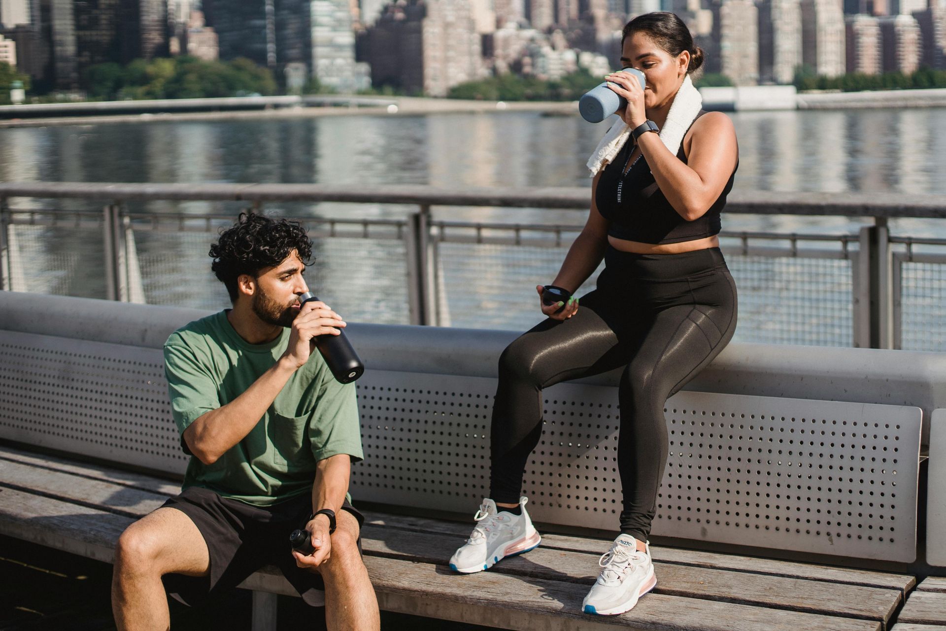 Two people resting after exercising, drinking from water bottles near a waterfront.