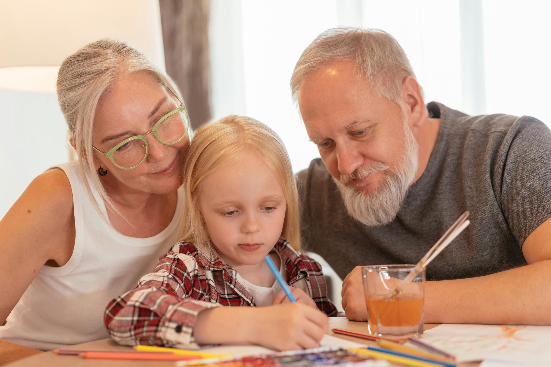 Grandparents watch a child draw with colored pencils at a table.