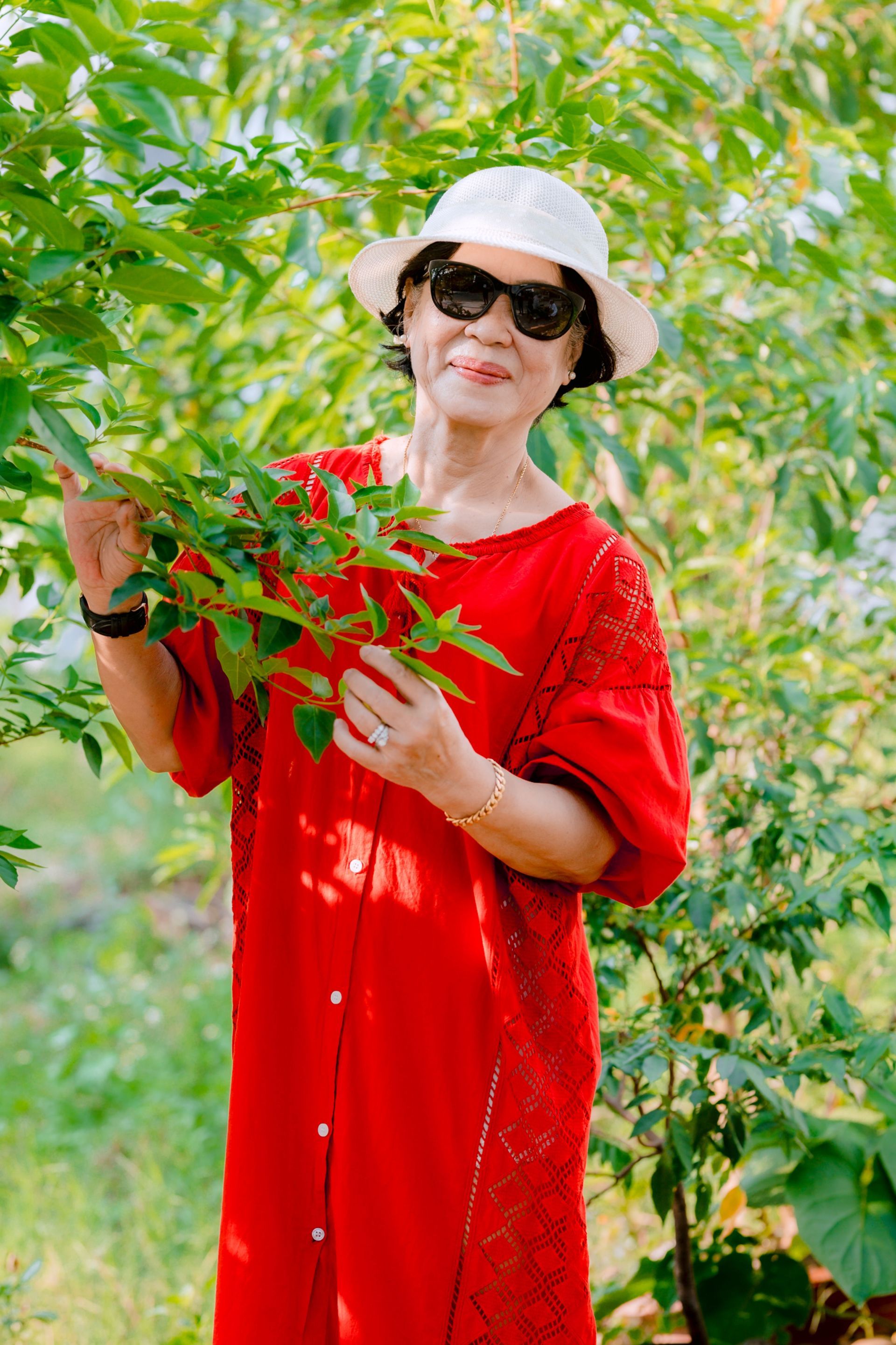 Woman in a red dress and white sunhat holding red flowers in a lush green garden.