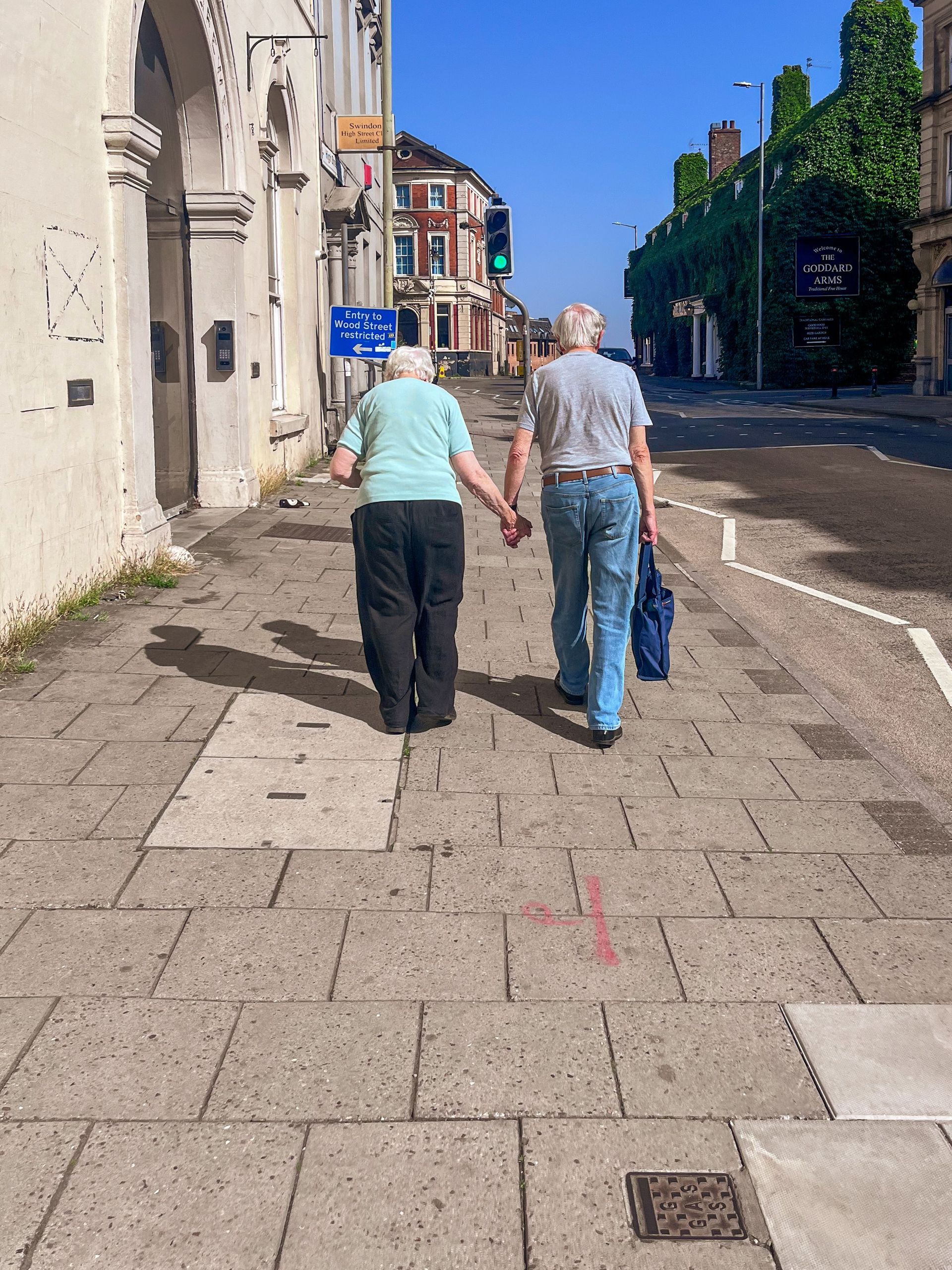 Two people walking hand in hand on a sunny city sidewalk beside buildings and trees.
