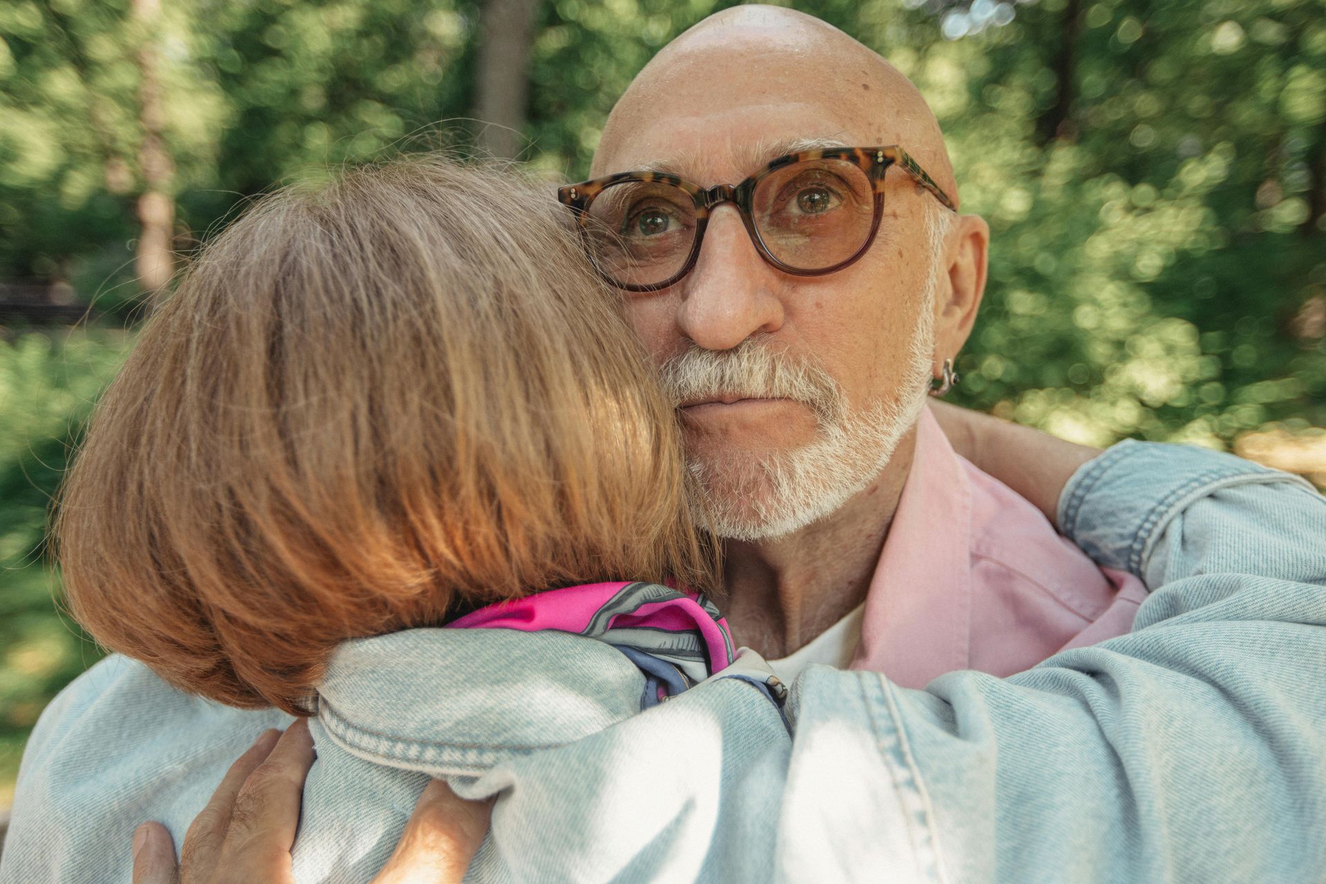 Bald person in glasses hugs someone. Outdoors with trees in the background.