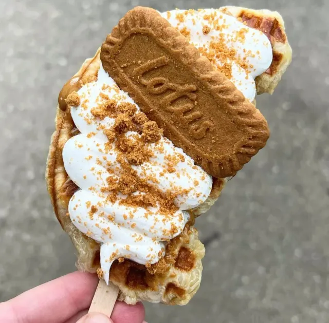 Handheld ice cream cone topped with whipped cream, crushed cookie crumbs, and a Biscoff cookie.