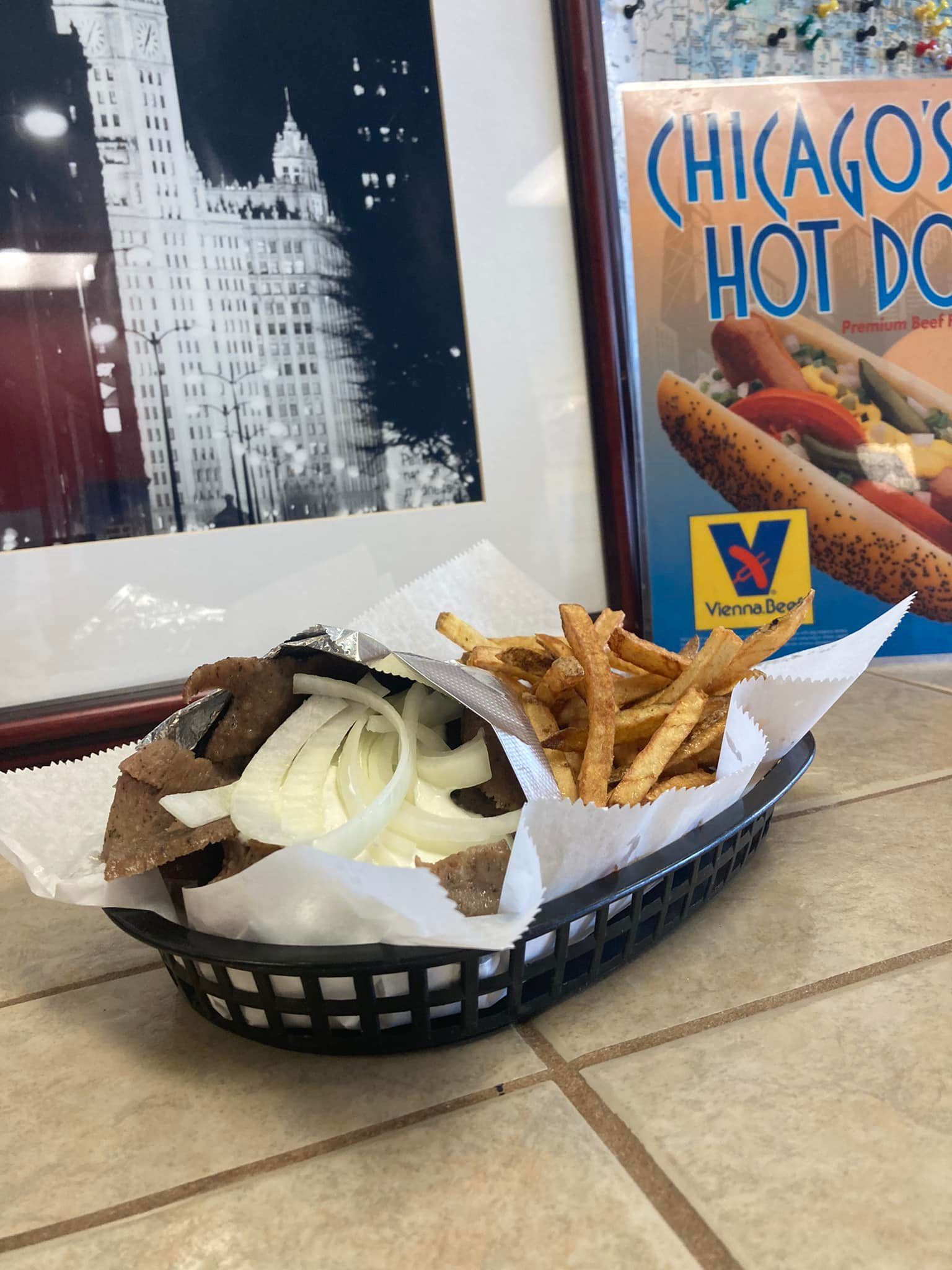Basket of Chicago-style hot dogs and fries on a table beside a Chicago-themed wall sign.