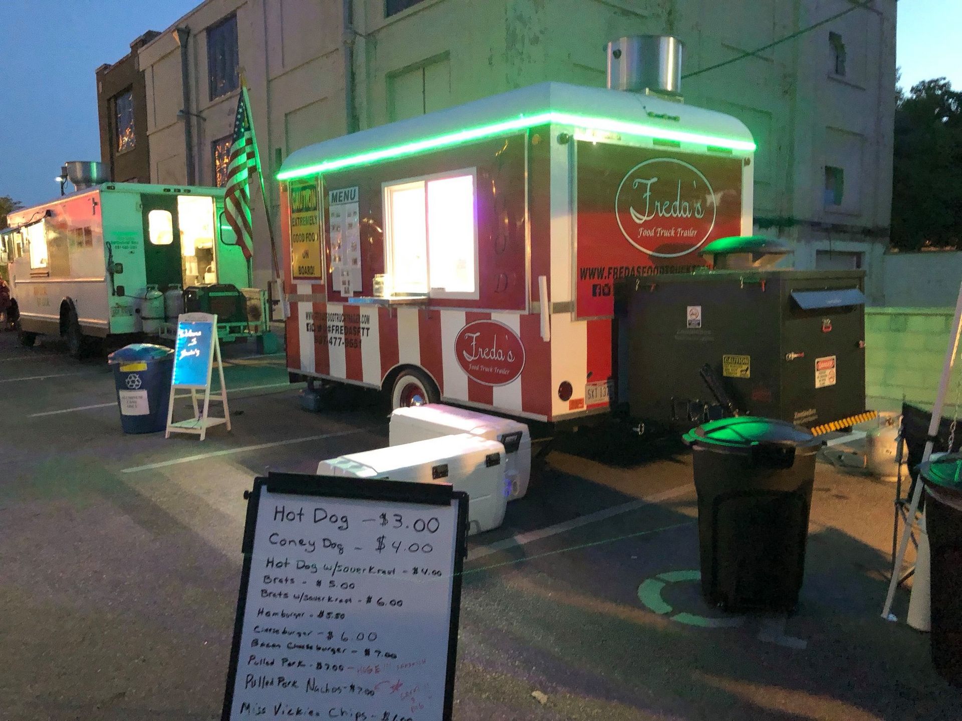 Food truck with neon green lights parked on a street at dusk, with a menu board and trash bins nearby.