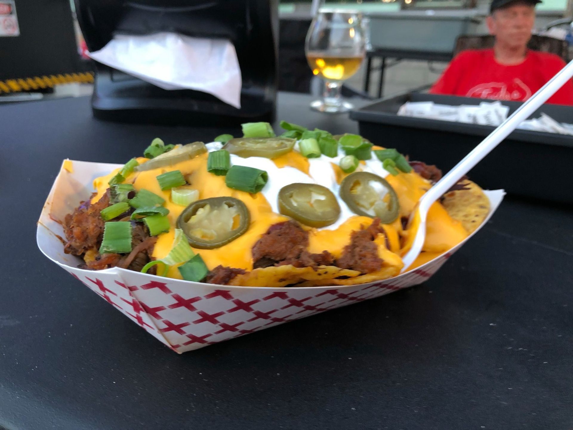 Loaded nachos with cheese, jalapeños, sour cream, and green onions in a takeout tray on a table.