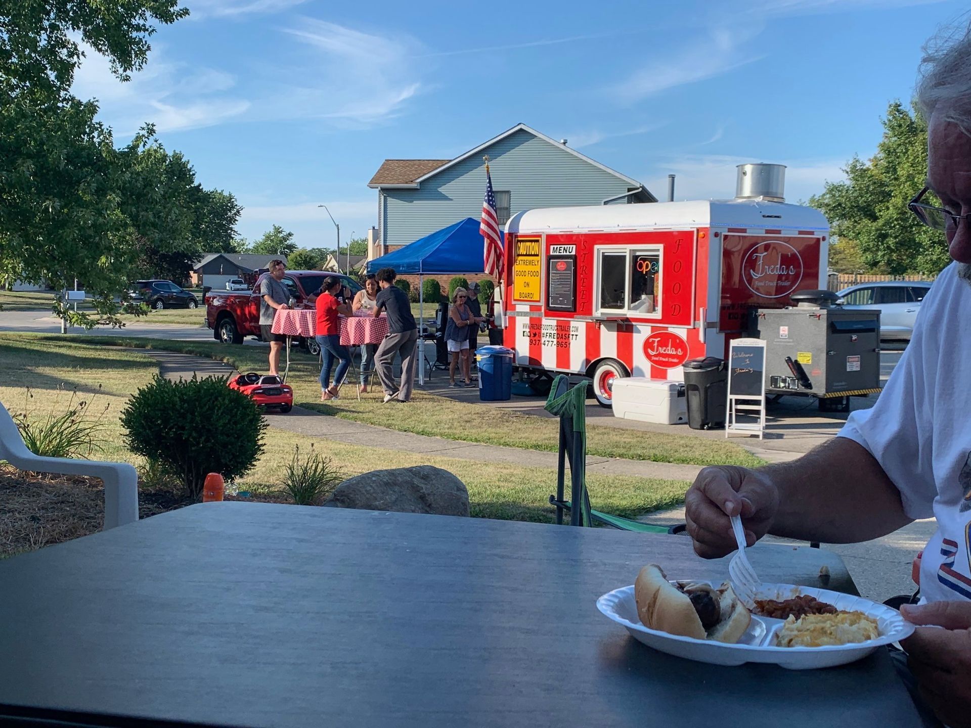 People gathered outside a red food truck near a waterfront, with a person eating in the foreground.