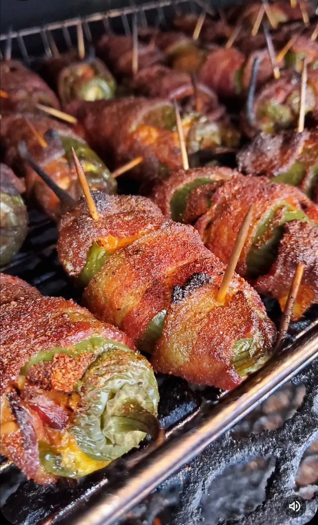 Grilled stuffed vegetables wrapped in bacon, sizzling on a metal rack over charcoal.