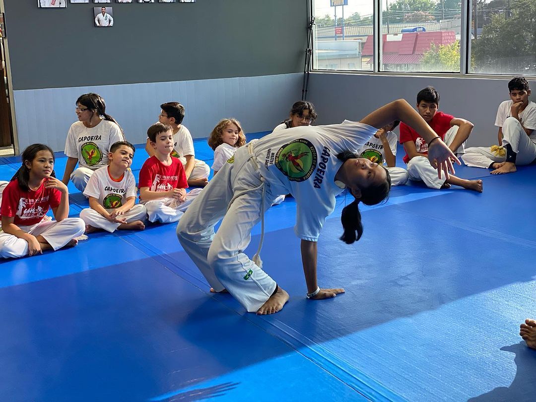 a group of children are sitting on the floor in a gym watching a woman perform capoeira .