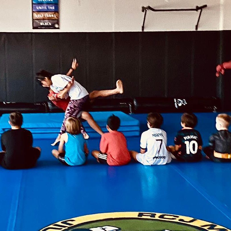 a group of kids are sitting on a blue mat in a gym and one of them has the number 10 on his back