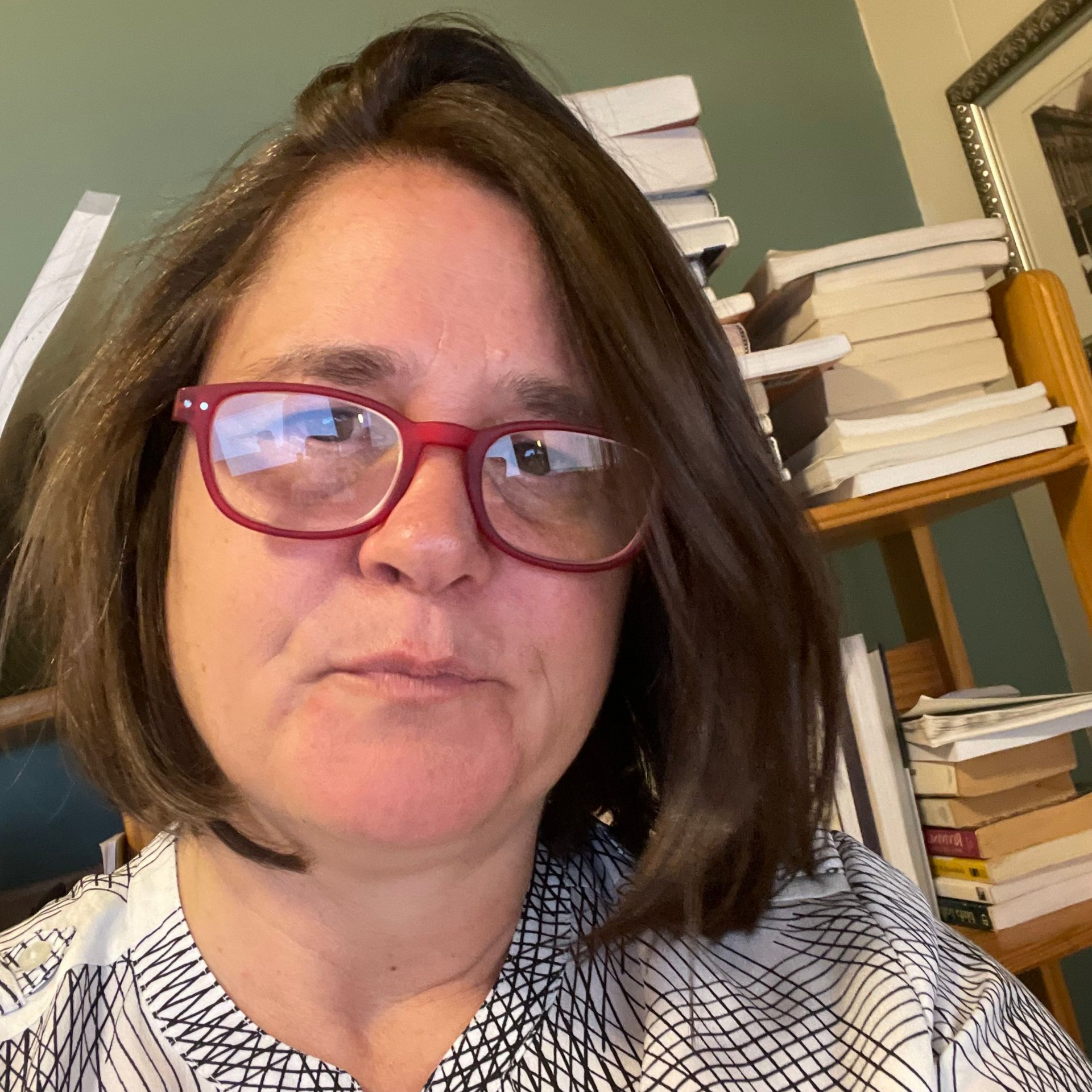 Woman with glasses and a patterned shirt, looks at the camera, surrounded by books.