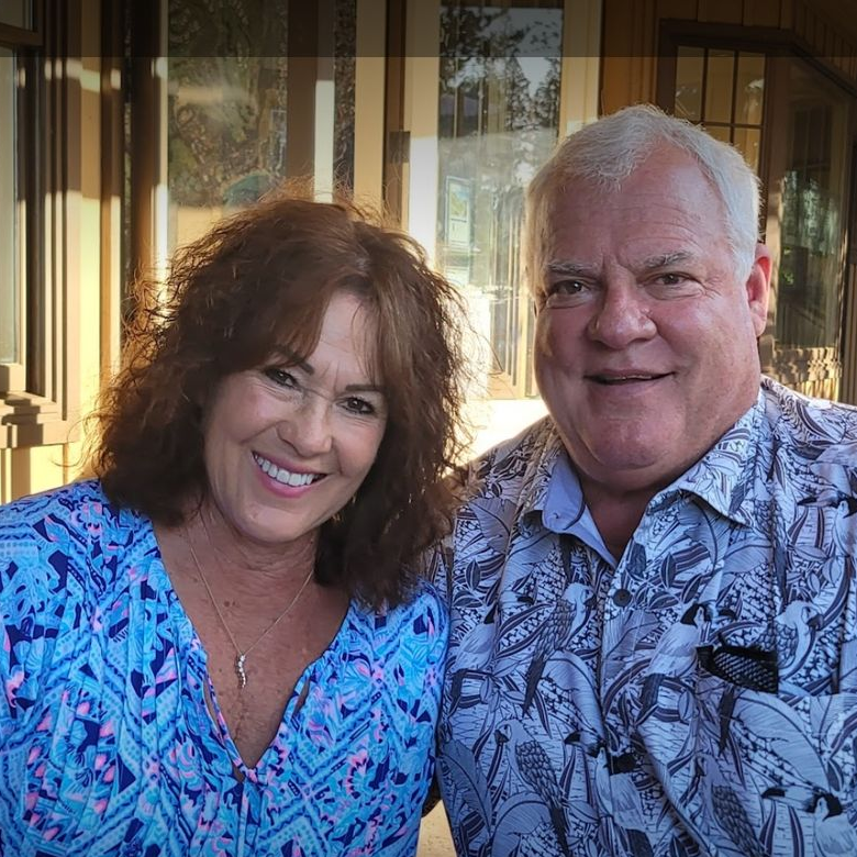 Smiling couple sitting on a couch, woman in blue dress, man in Hawaiian shirt, outdoor setting.
