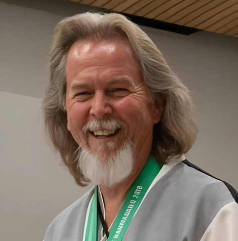 Doug Fuechsel headshot: Smiling man with gray hair and beard wearing a gray and white uniform, medal around his neck.
