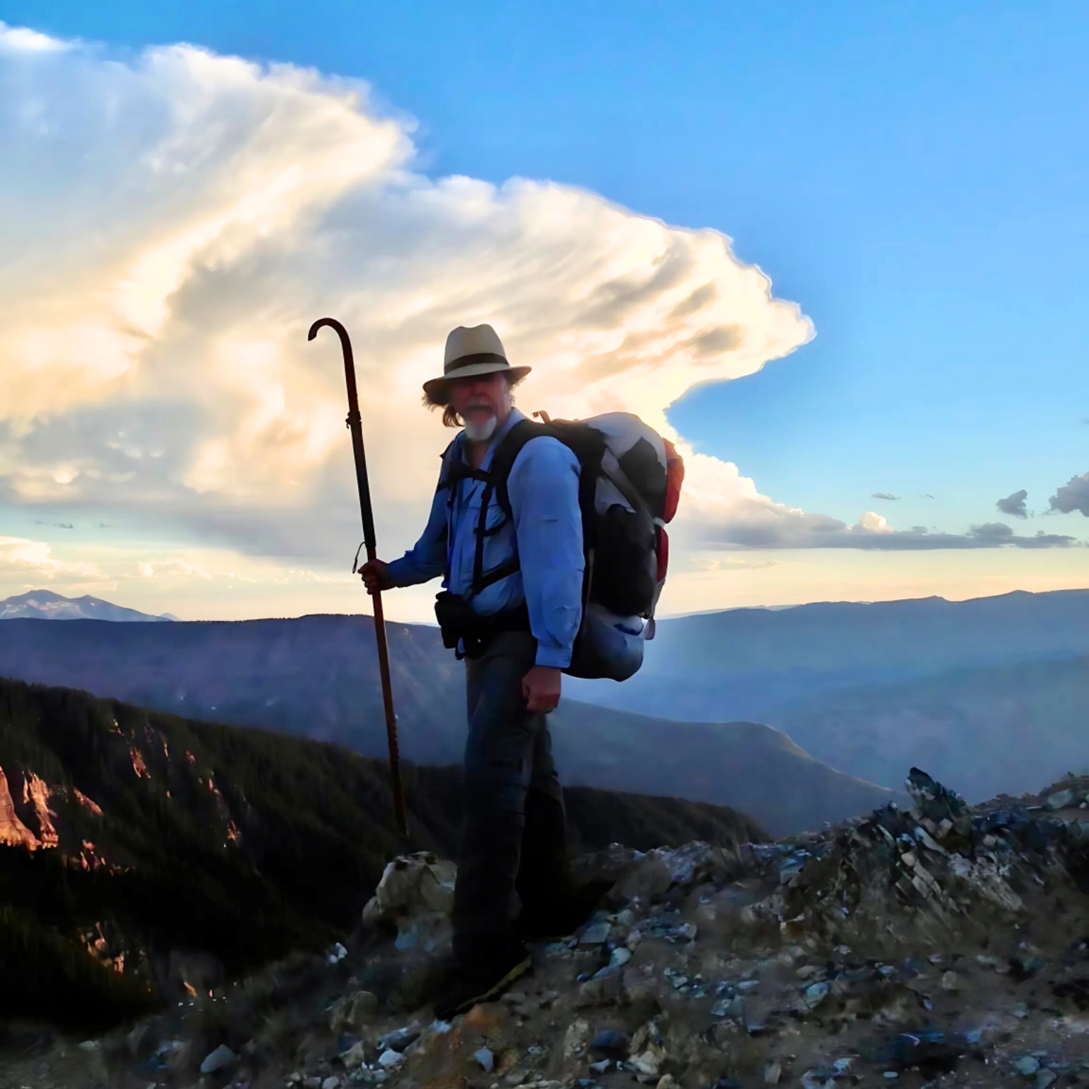 Hiker in a hat, with a backpack and walking stick, stands on a mountain ridge, under a dramatic cloud.