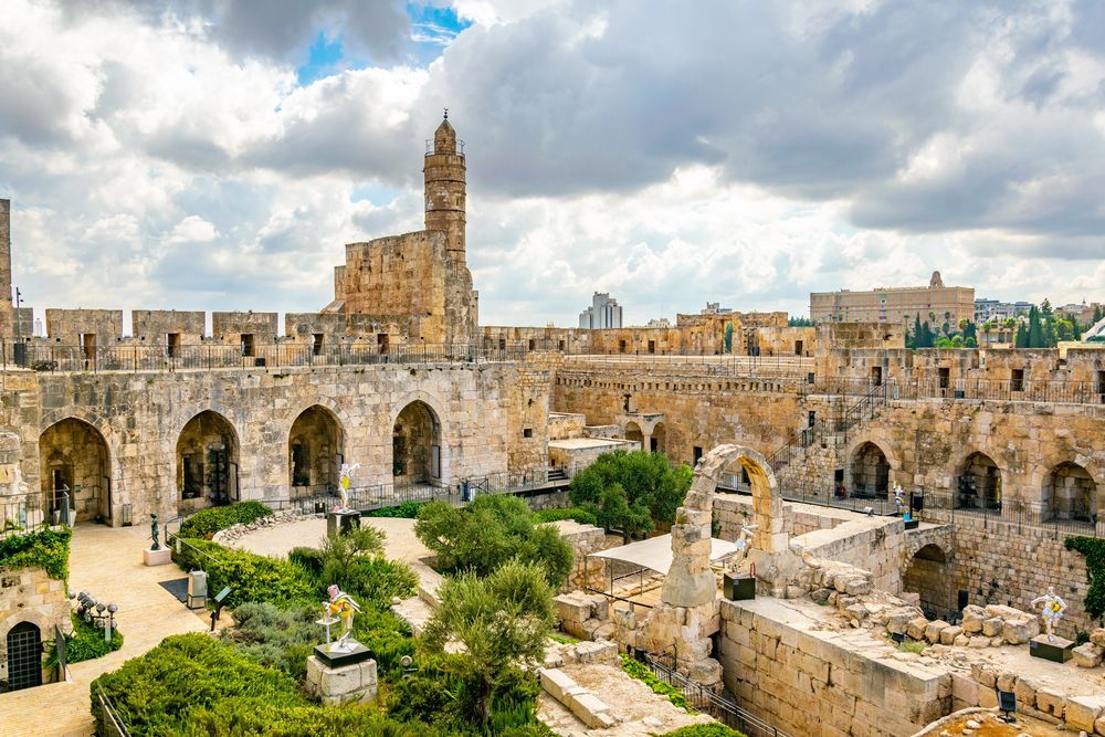 Jerusalem citadel: ancient stone walls, tower, arches, courtyard, cloudy sky, greenery, buildings.