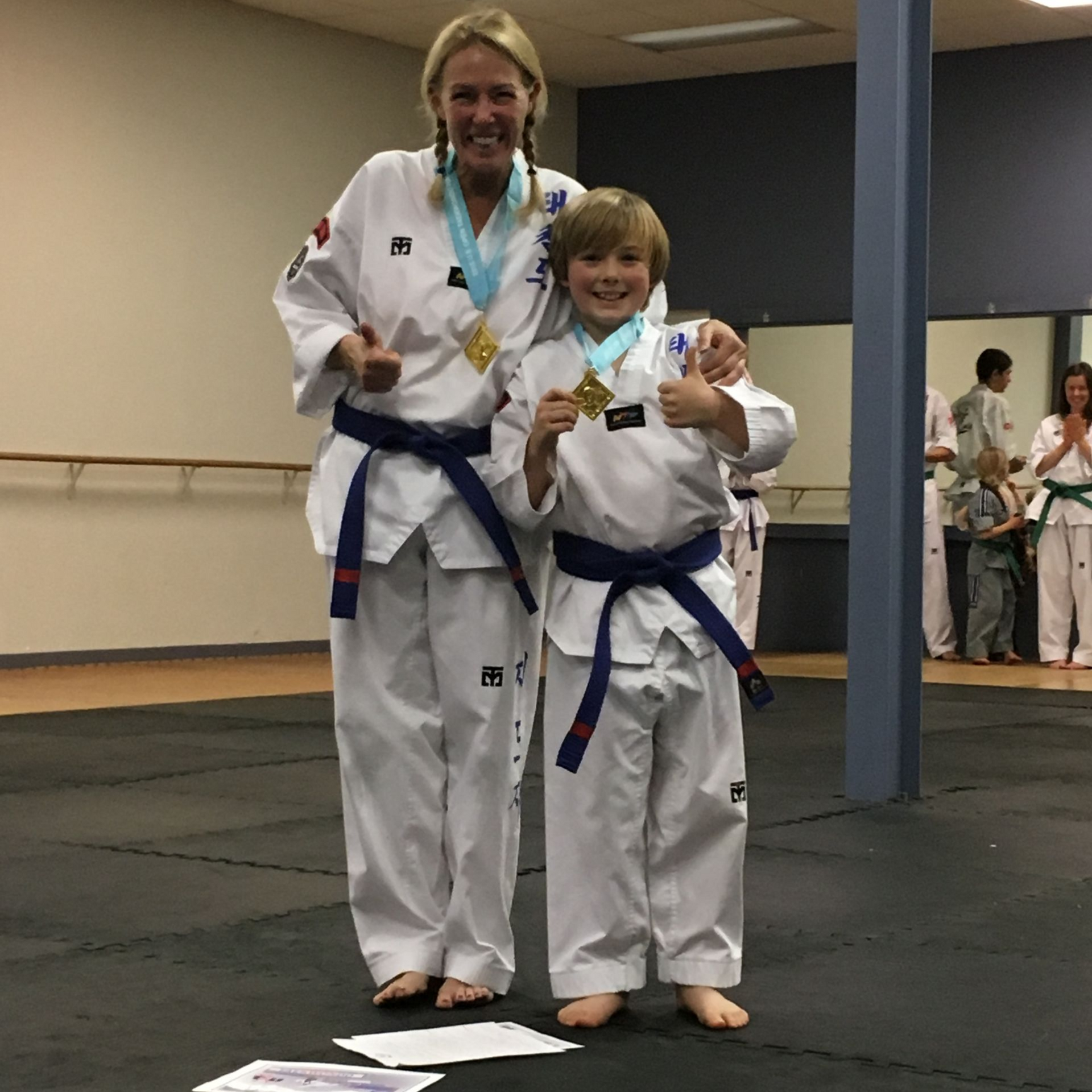 Woman and boy in martial arts uniforms with gold medals, giving thumbs up. Indoor setting.