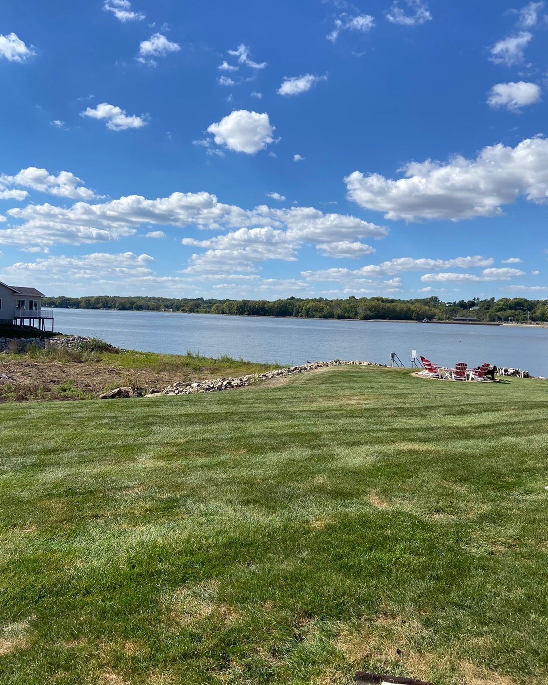 Lush green lawn leading to a calm river under a blue sky with fluffy white clouds.