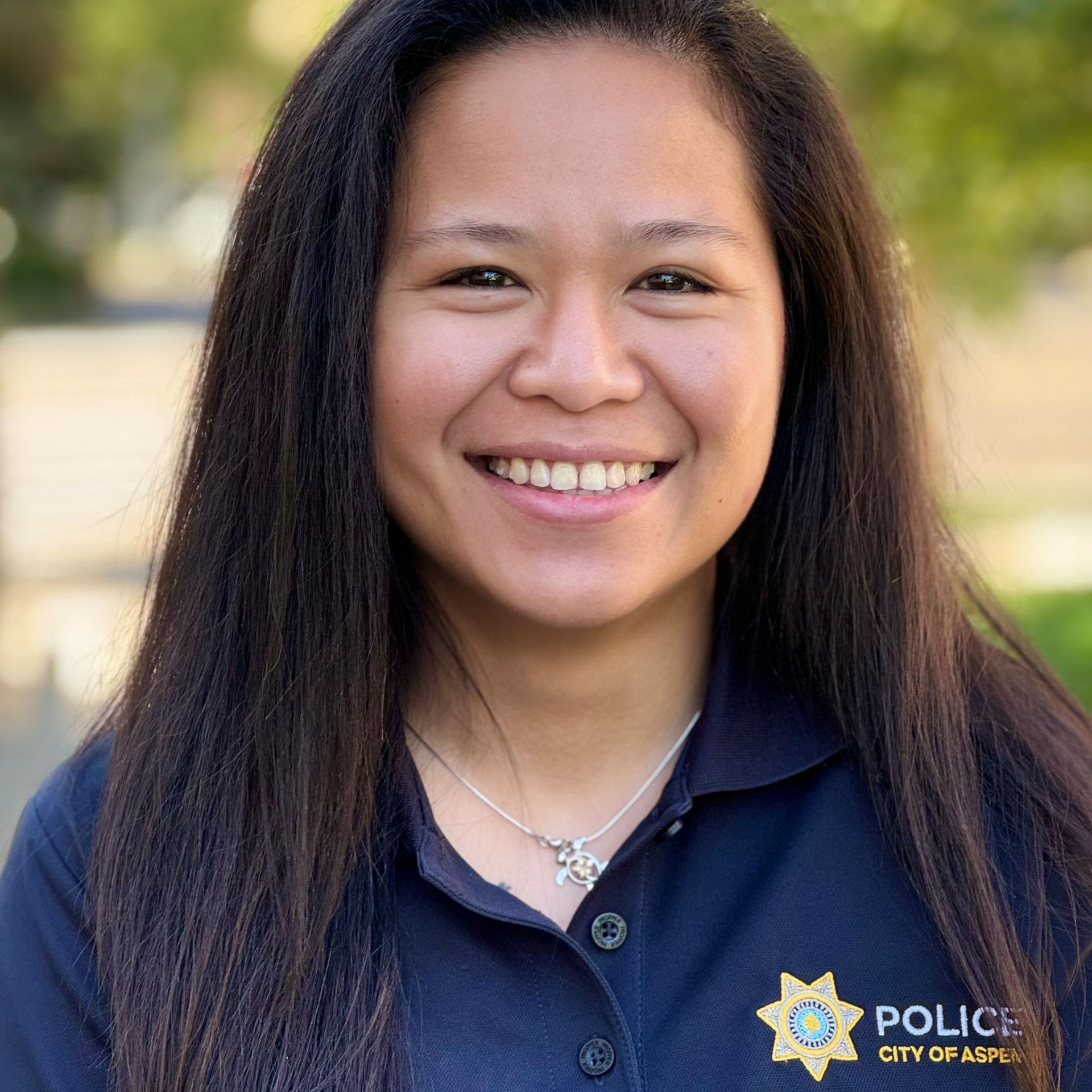 Woman with long dark hair smiles at the camera, wearing a navy blue police uniform shirt, outdoors.