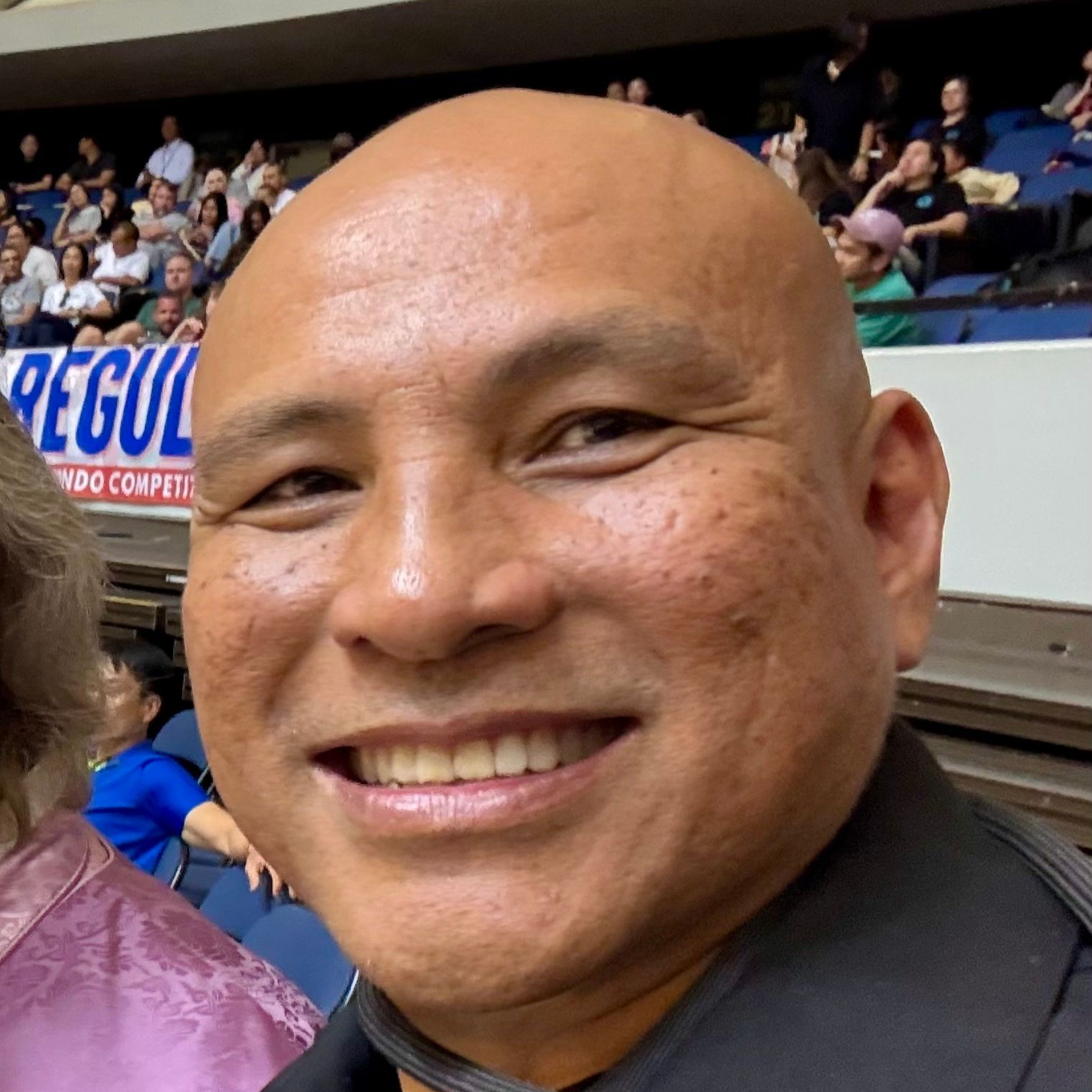 Smiling bald man, dark complexion, wearing a black shirt; arena seating in the background.