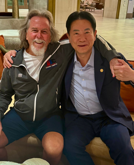 Two men seated, smiling, arm around each other. One with long gray hair, the other in a suit. Indoor setting.