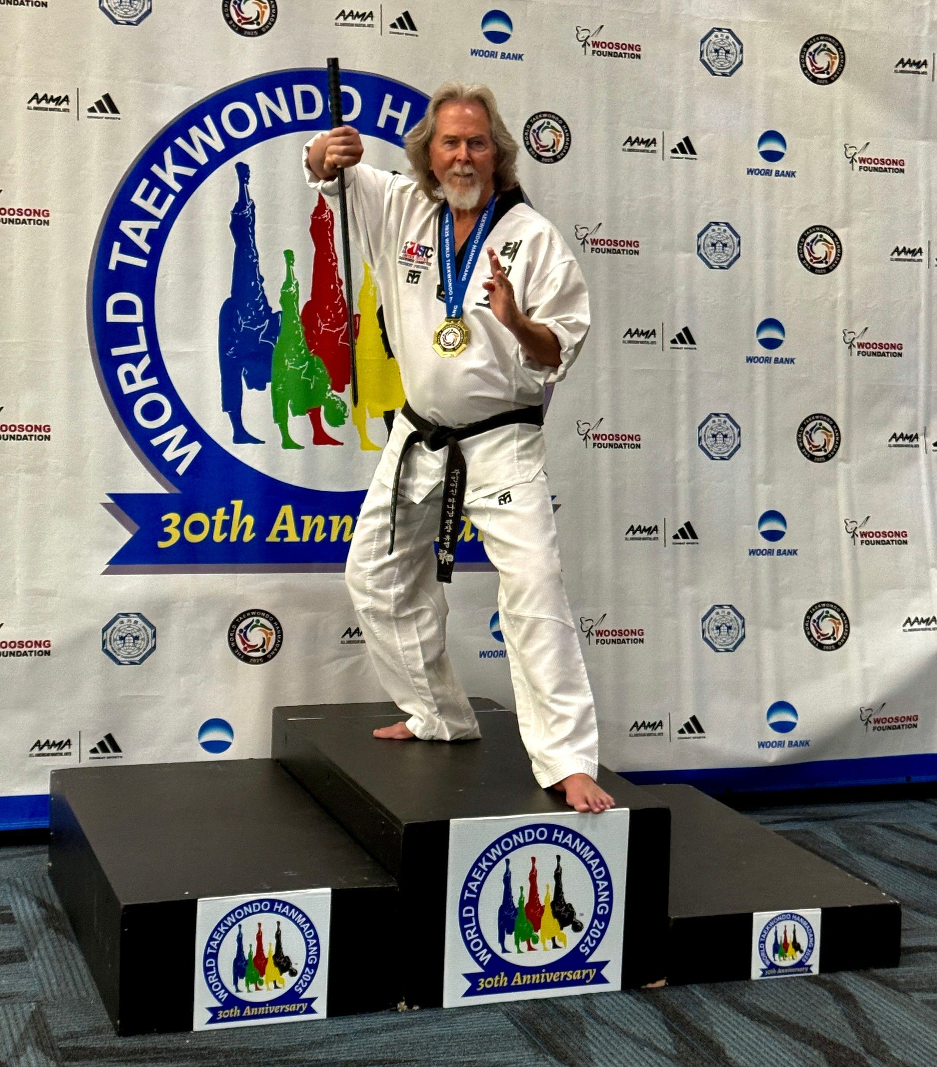 Man in black belt taekwondo uniform, gold medal, on podium at World Taekwondo Championship, holding up a staff.