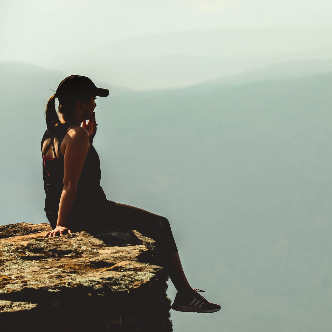 Woman in athletic wear sits on a cliff, looking out at a distant mountain range.