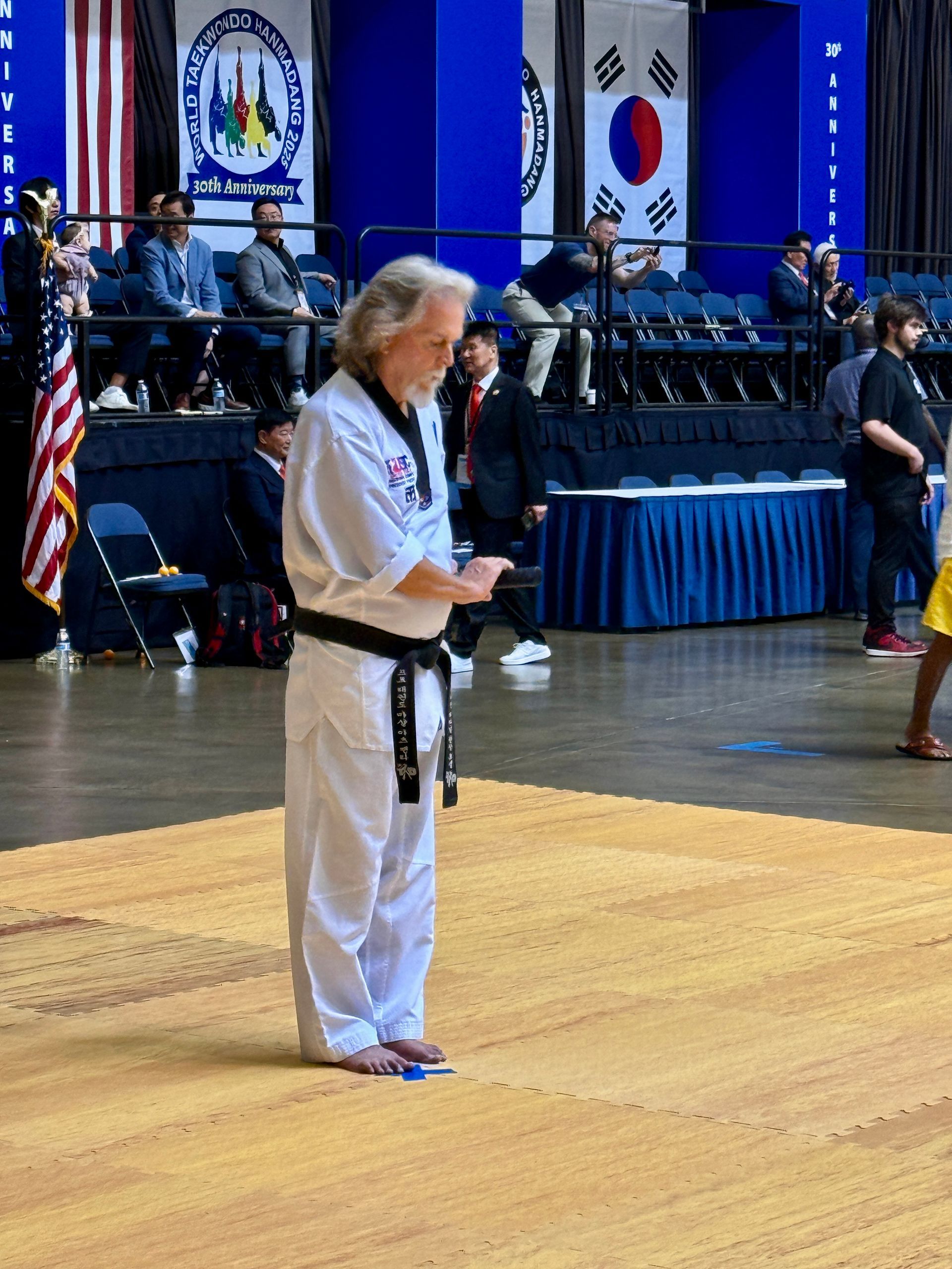 Man in white martial arts uniform, black belt, bowing on a mat at a competition.