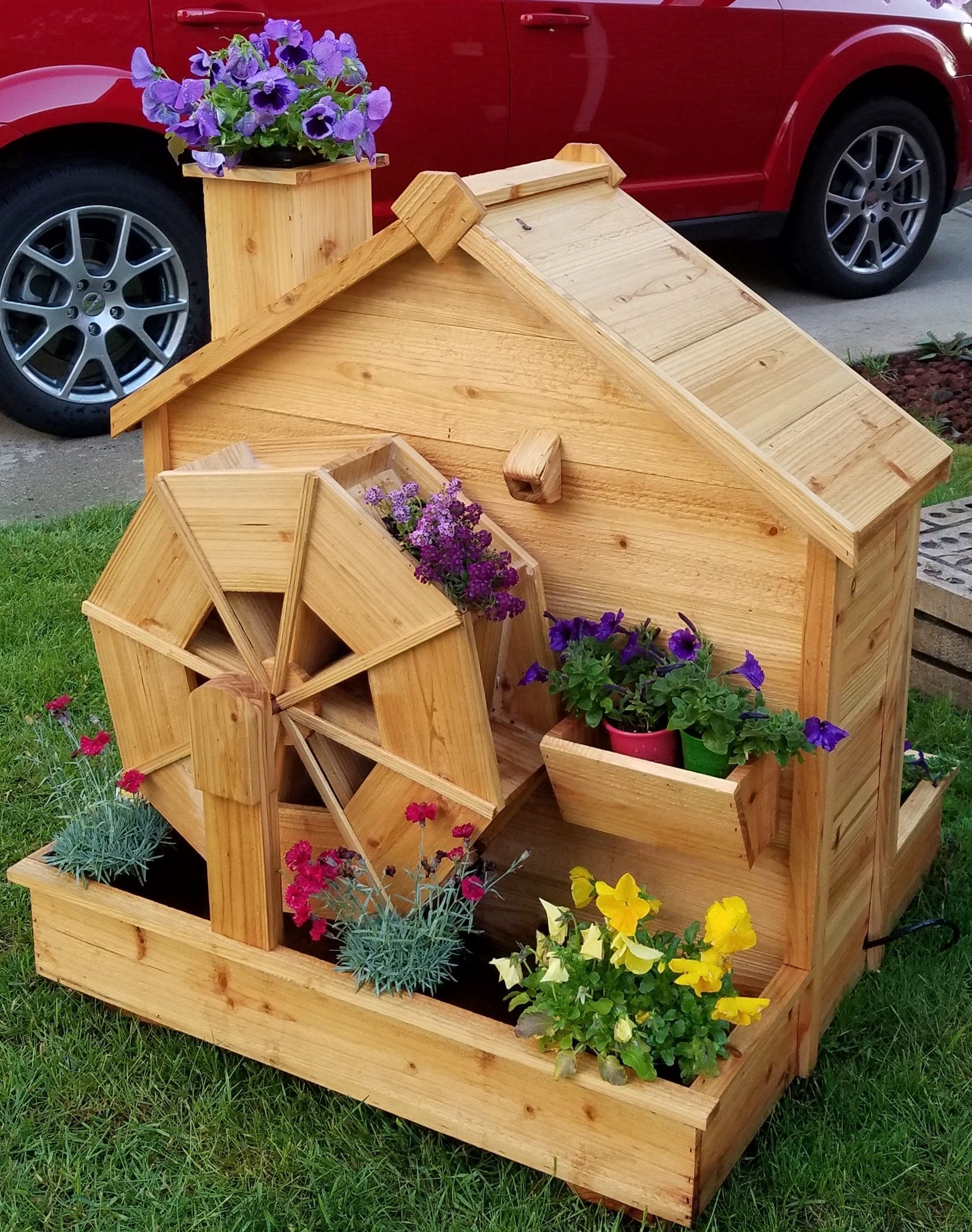 A wooden water wheel with flowers in front of a red car.