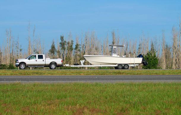 White truck towing a boat trailer with a white boat on a highway with green grass and trees.